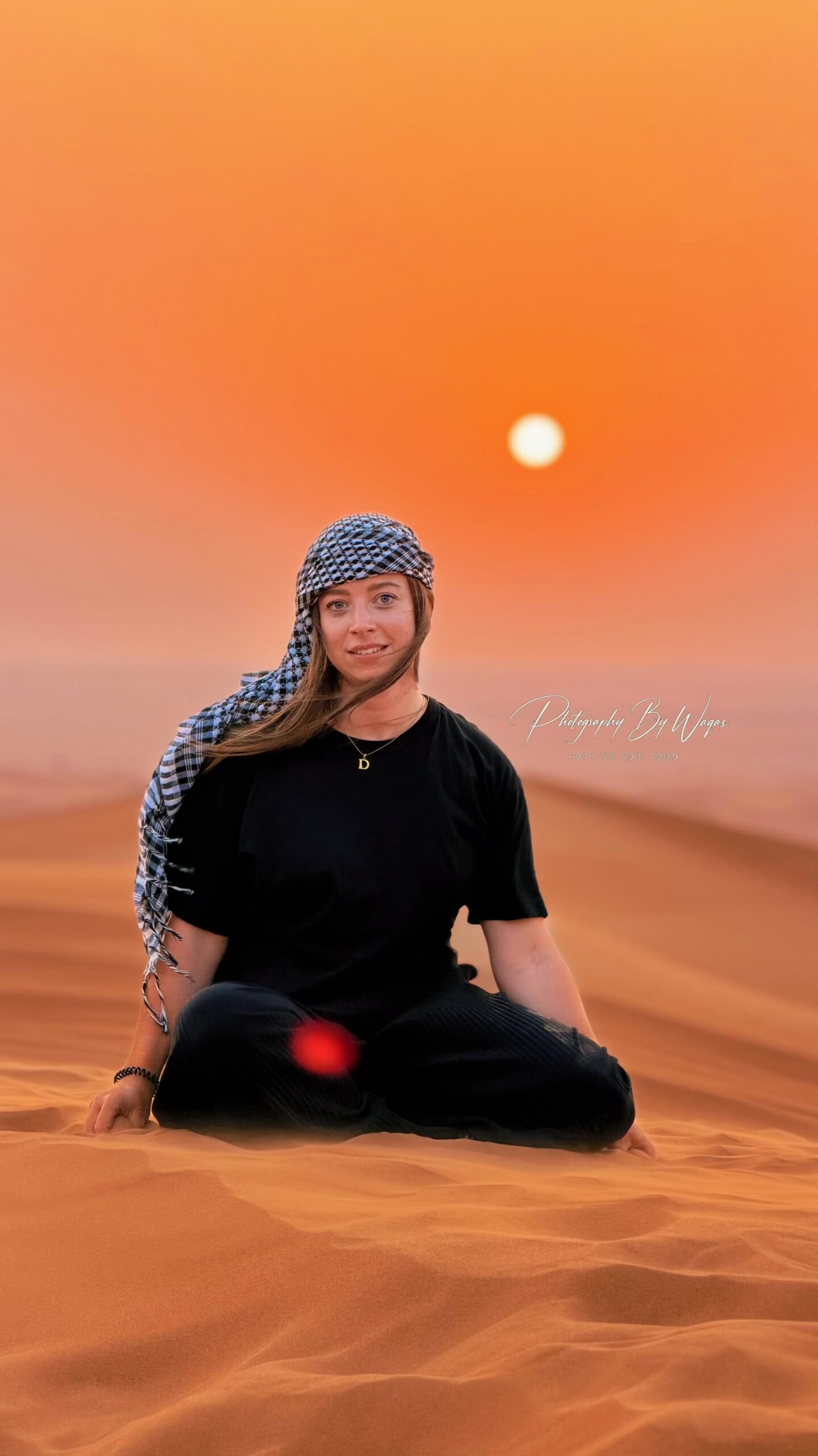 Tourist woman wearing a black t-shirt, pants, and a patterned shemagh headscarf meditating in a cross-legged pose on the red sand dunes during a desert sunset photoshoot in Dubai